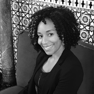 Black and white photo of Barika Williams. She has her head turned to the camera and is smiling. A tiled wall is behind her.